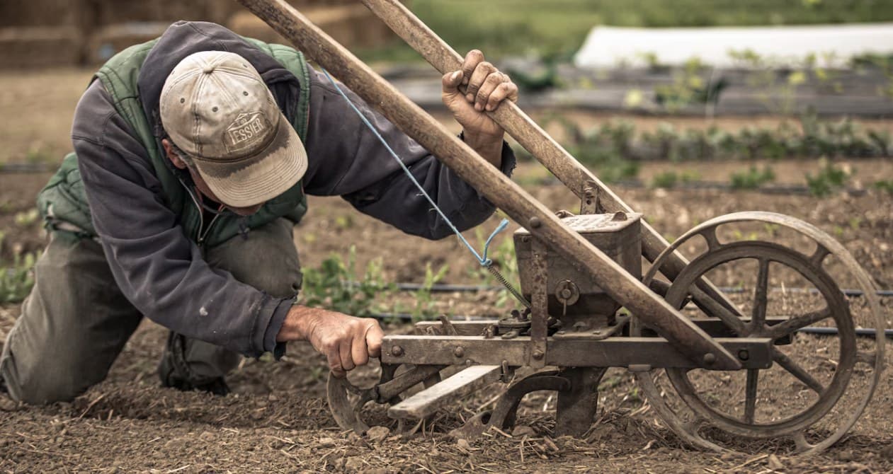 A farmer on his knees adjusting a hand seeder in a planted row